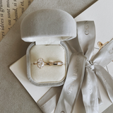 A pair of gold rings, including an oval diamond engagement ring and a matching band, displayed in a gray velvet ring box with a champagne ribbon from Concept26, set on a beige textured surface with book pages.